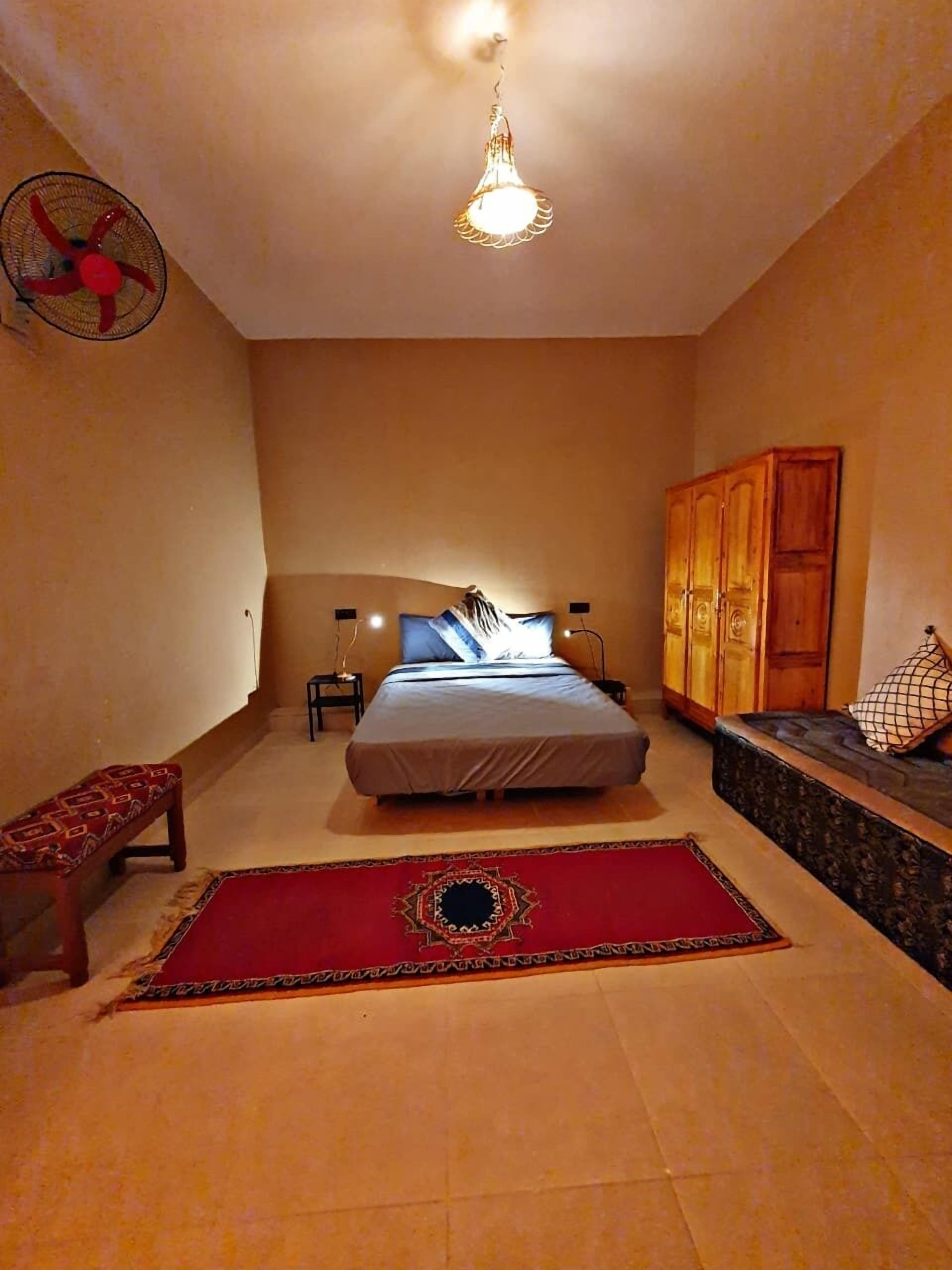 Ein Schlafzimmer mit einem Bett, einem Holzschrank, einer Bank, einem roten Teppich mit Muster und einem Deckenventilator.