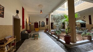 A spacious indoor area with a tiled floor, a sofa, a small table, and a bookshelf. There are potted plants and a guitar in the room. - Chambres d'Hôtes Riad Dar Tazoulte (Agdz)