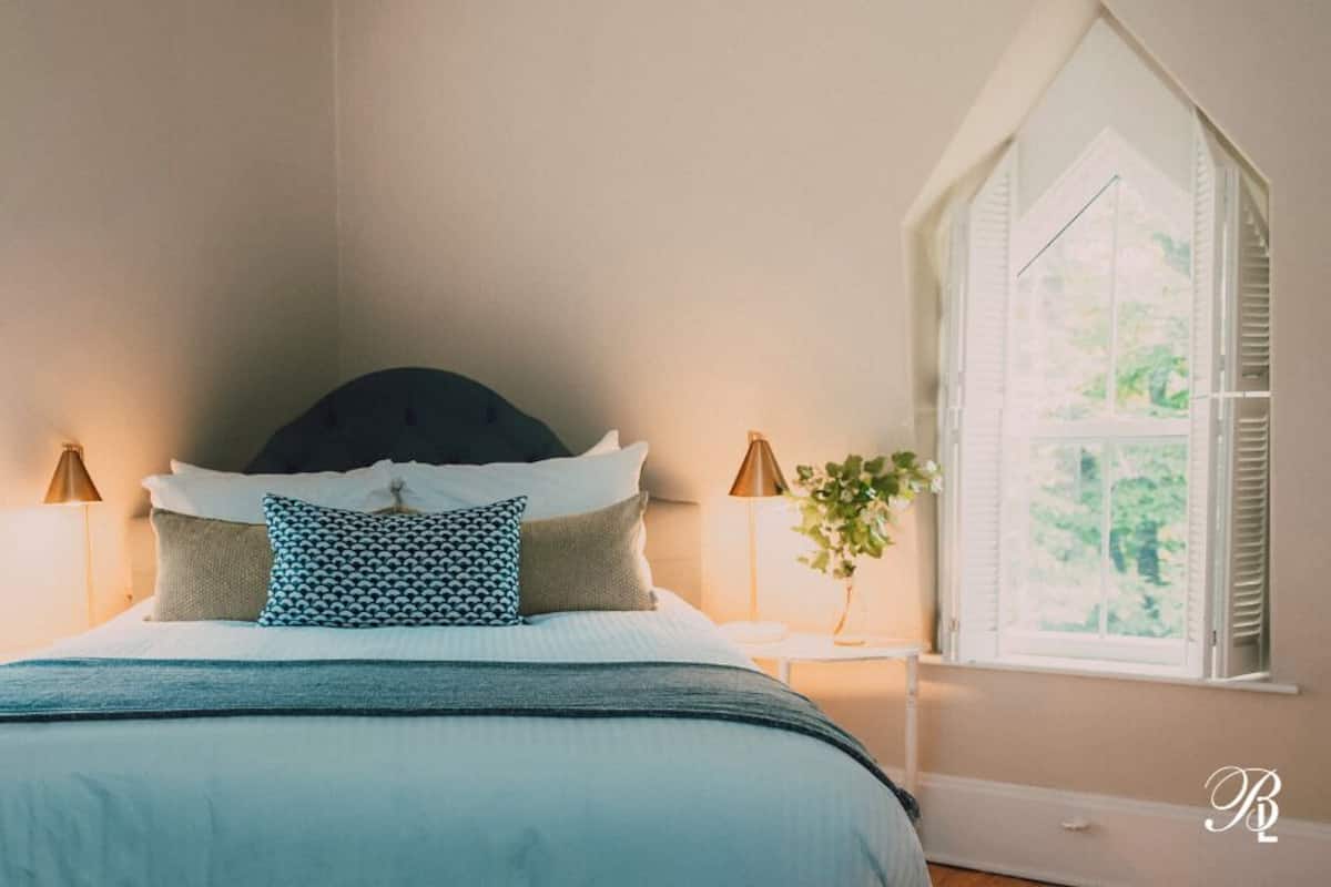 A bedroom with a bed, two bedside lamps, a window with shutters, and a plant on the nightstand.