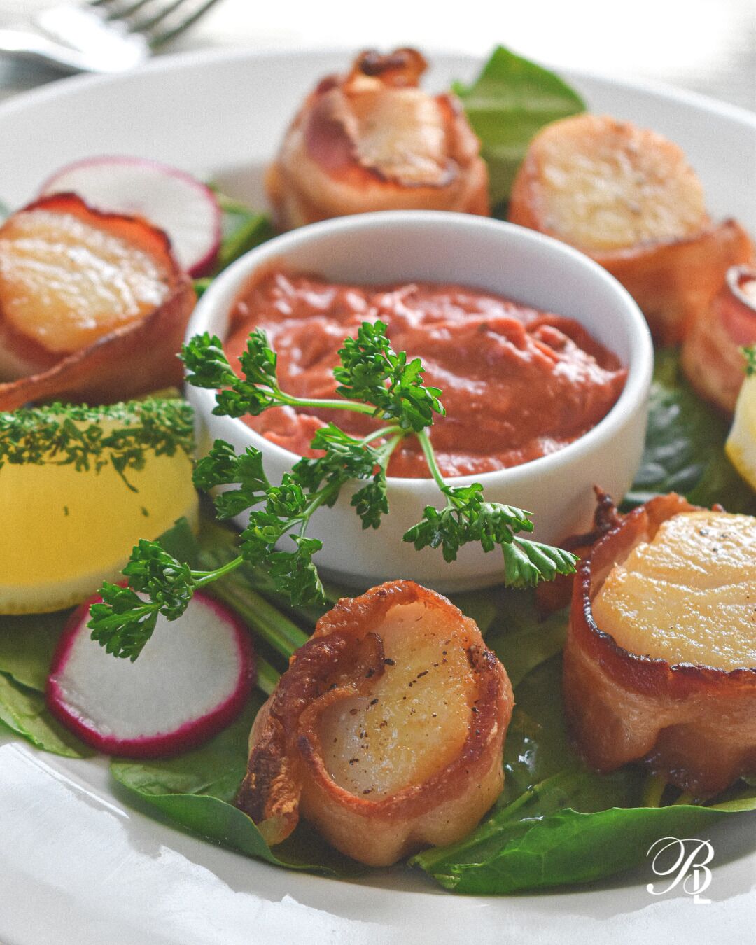 Un plato con diversos alimentos, incluyendo vieiras envueltas en tocino, rábanos y un recipiente con salsa, adornado con perejil.