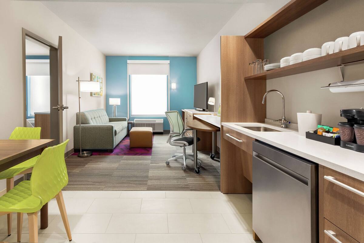 Une chambre d’hôtel moderne dotée d’un coin repas, d’un canapé, d’un bureau et d’un coin cuisine avec un lave-vaisselle et des étagères.