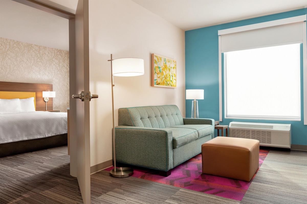 A hotel room with a bed, a sofa, a chair, a standing lamp, a painting, a window, and an air conditioning unit.