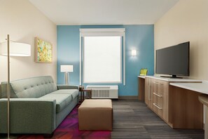 A modern hotel room with a sofa, a flat-screen TV, a desk, a lamp, and a window.