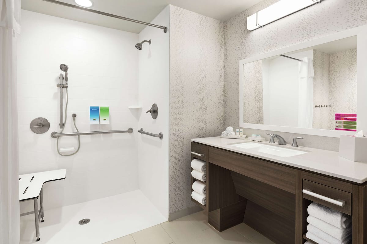 Une salle de bain moderne dotée d’une douche à l’italienne, d’un meuble-lavabo avec un grand miroir et de toilettes équipées d’une barre d’appui.