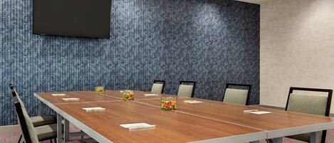 A conference room with a long table, six chairs, a flat-screen TV mounted on the wall, and a patterned carpet.