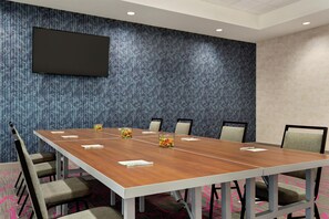 Une salle de conférence avec une longue table, six chaises, un téléviseur à écran plat fixé au mur et une moquette à motifs.