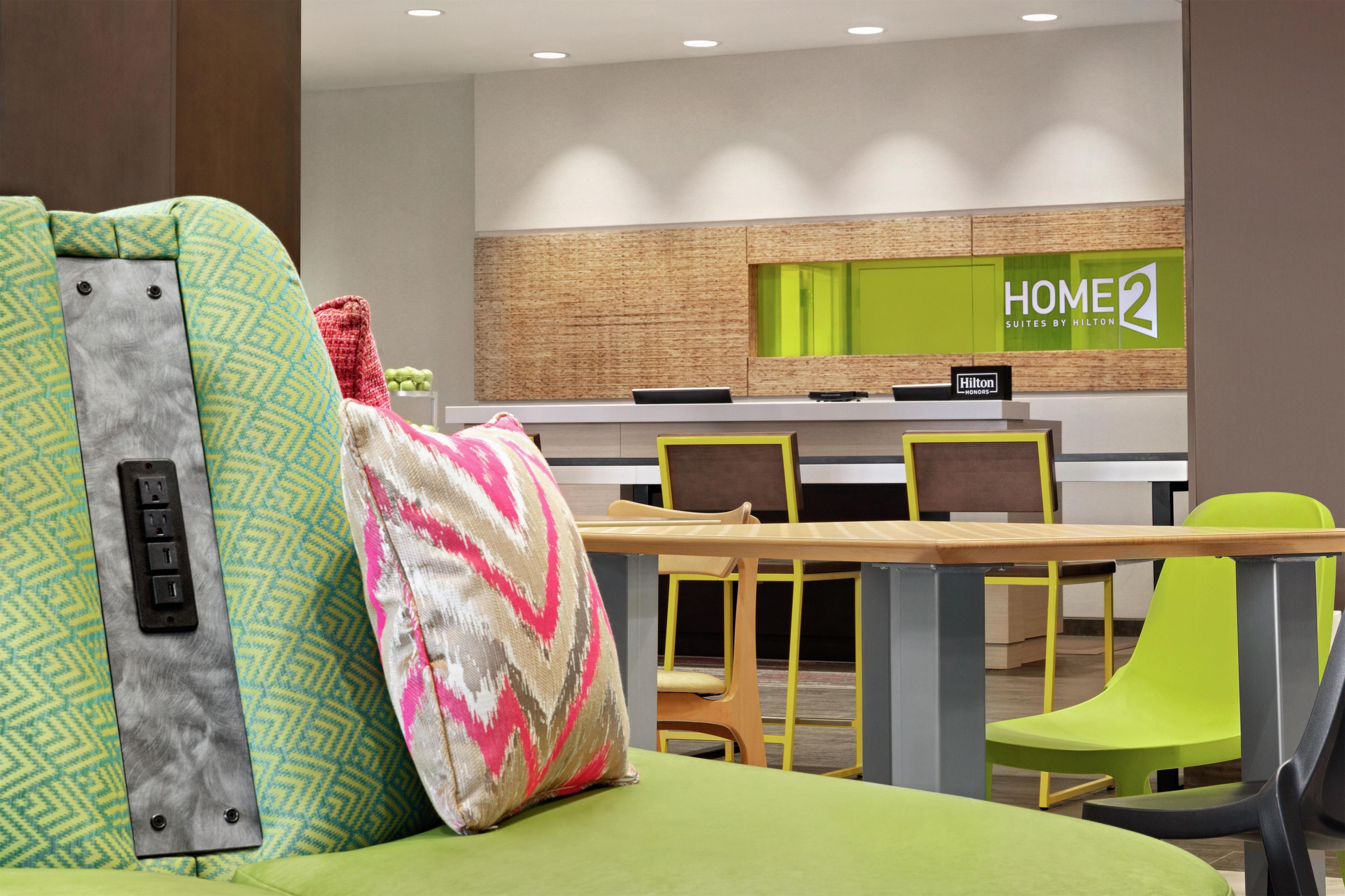 Une chambre d’hôtel avec un lit, un bureau et des chaises. Il y a une enseigne sur le mur qui indique : « HOME2 SUITES BY HILTON ».