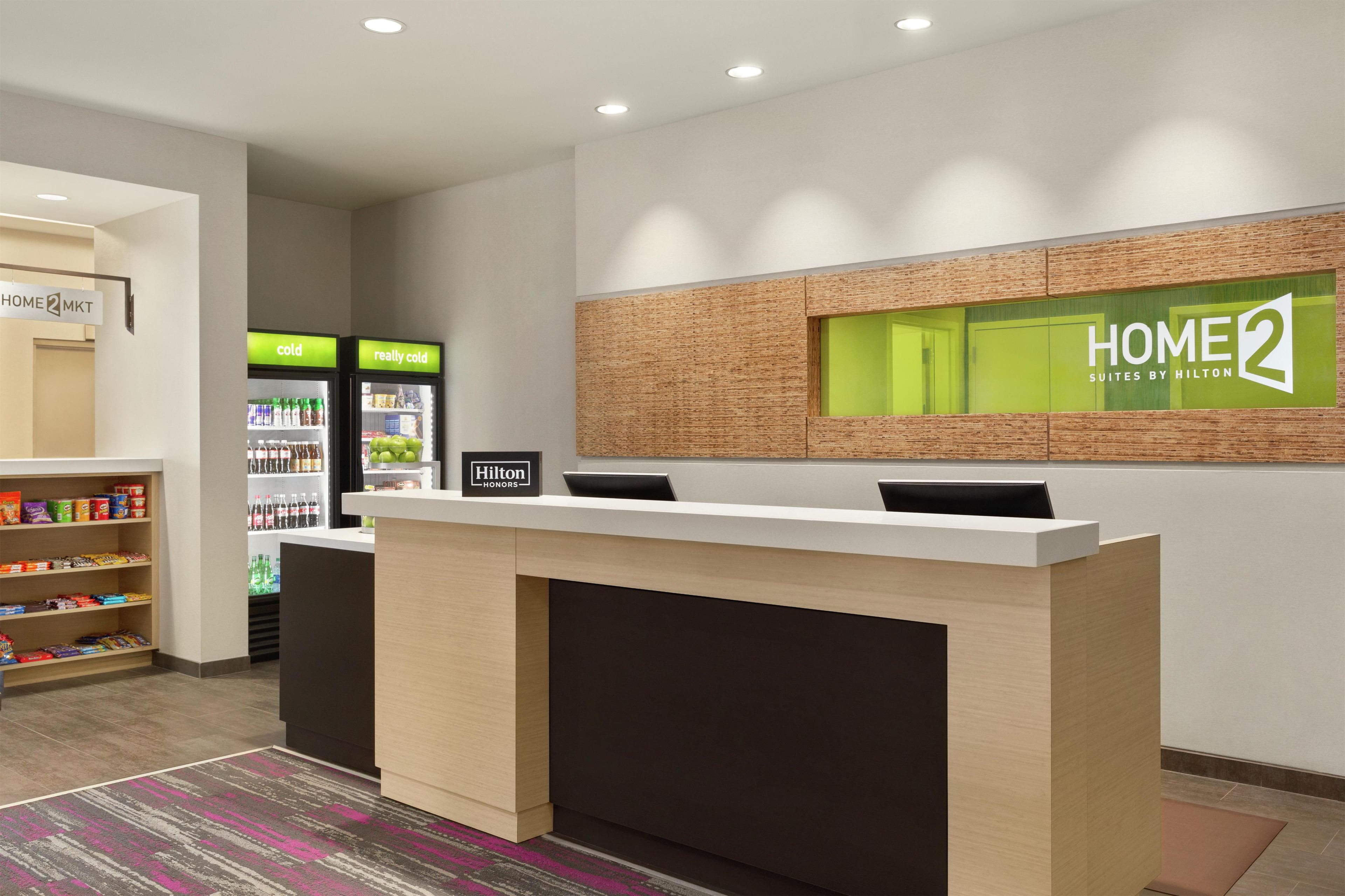 A hotel reception desk with a 'HOME 2 SUITES BY HILTON' sign, a minibar, and a 'Hilton Honors' label.
