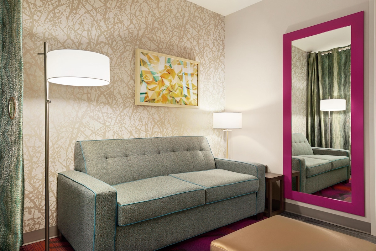 A hotel room with a sofa, a side table, a lamp, a mirror, and a patterned wall.