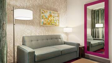 A hotel room with a sofa, a side table, a lamp, a mirror, and a patterned wall.