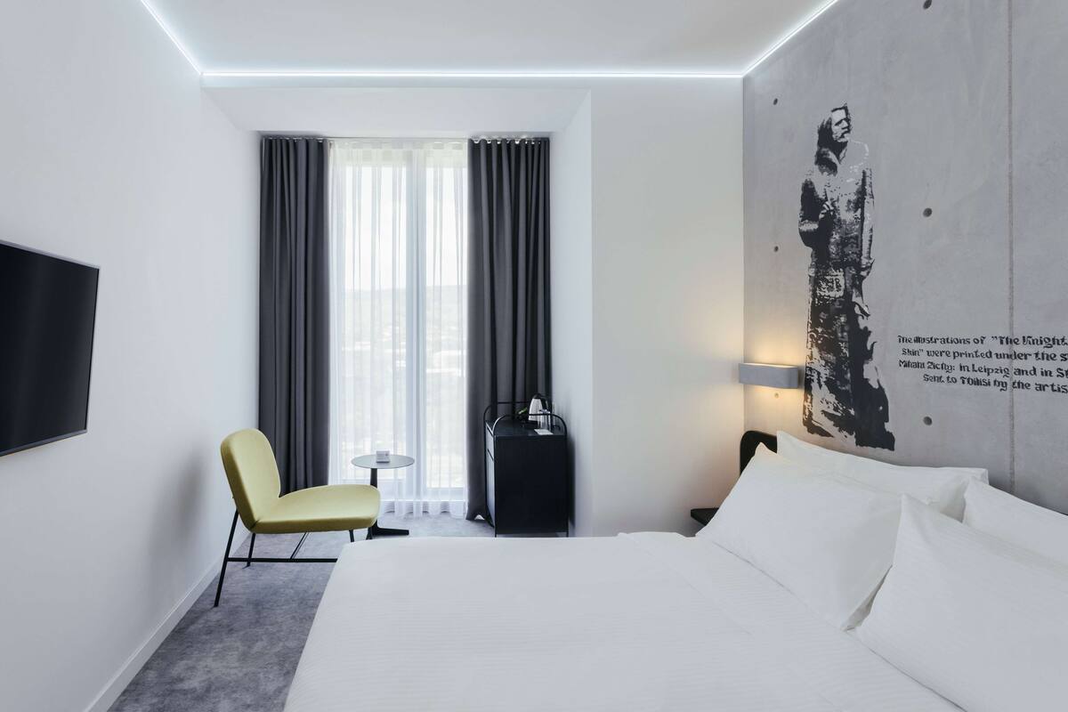 A modern hotel room with a large bed, a flat-screen TV mounted on the wall, a small round table, a yellow chair, and a window with curtains.
