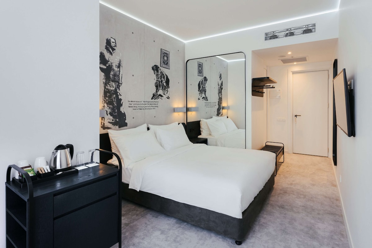 Une chambre à coucher moderne avec un grand lit, des tables de chevet, une commode noire, un miroir et des œuvres d’art accrochées aux murs.