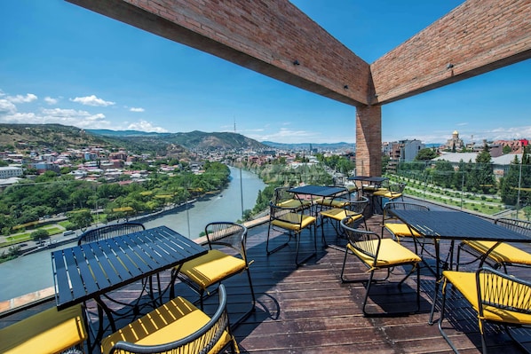 A terrace with outdoor seating overlooking a river and cityscape. - Golden Tulip Design Tbilisi (Tbilisi)