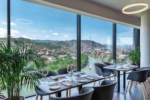 A dining area with a table set for two, large windows offering a view of a river and cityscape, and potted plants. - Golden Tulip Design Tbilisi (Tbilisi)
