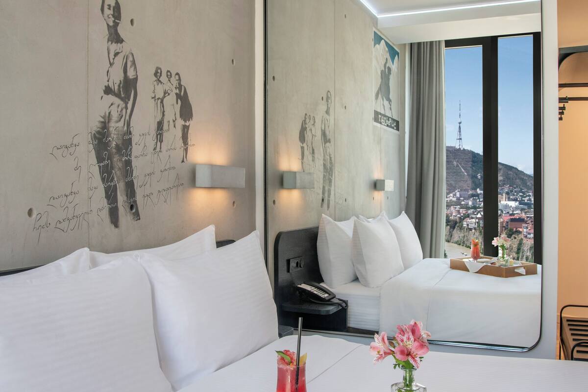 A hotel room with a large bed, a bedside table with a phone, a tray with food and drink, a mirror, and a city view.