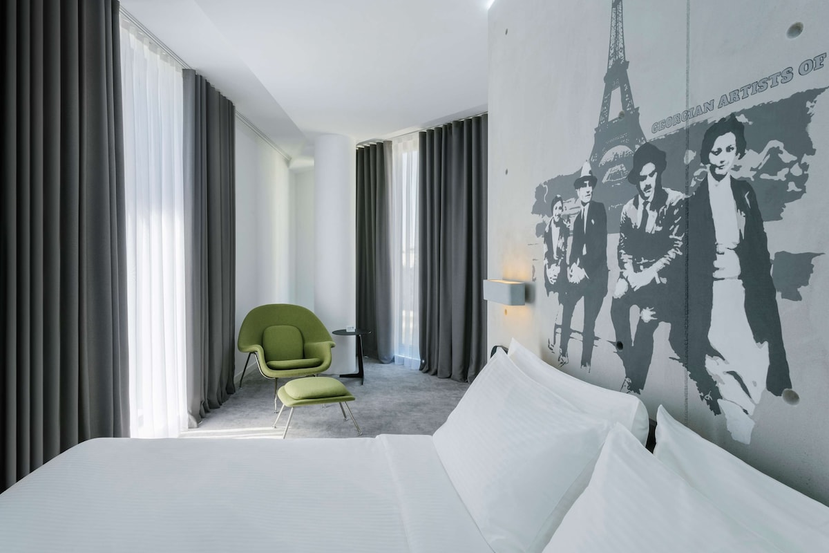 Une chambre d’hôtel moderne avec un grand lit, un fauteuil vert et une fresque murale représentant des œuvres d’artistes géorgiens.
