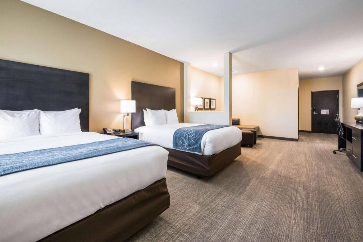 A hotel room with two beds, a desk, a chair, a lamp, and a door.