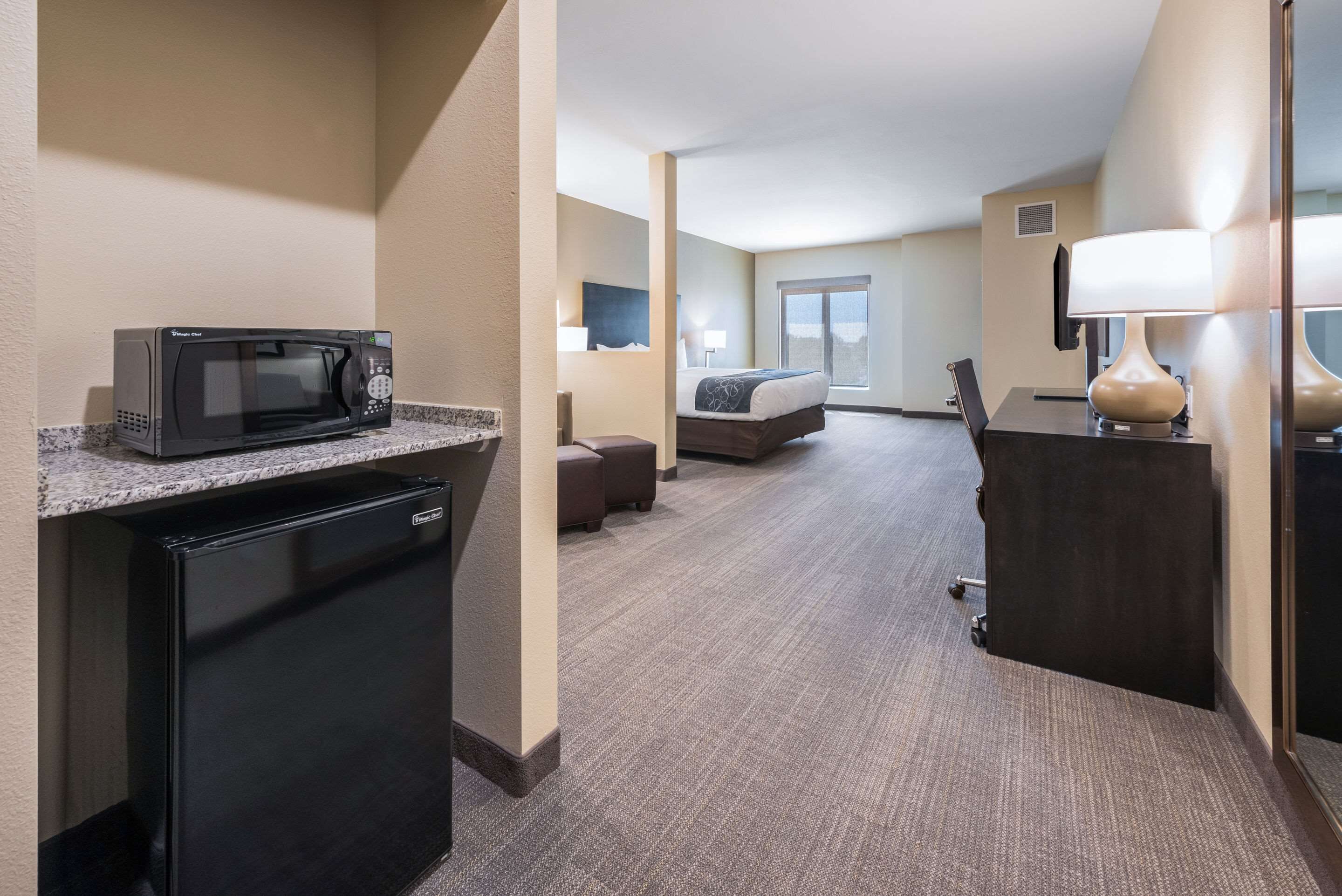 A hotel room with a kitchenette, a bed, a desk, and a lamp.
