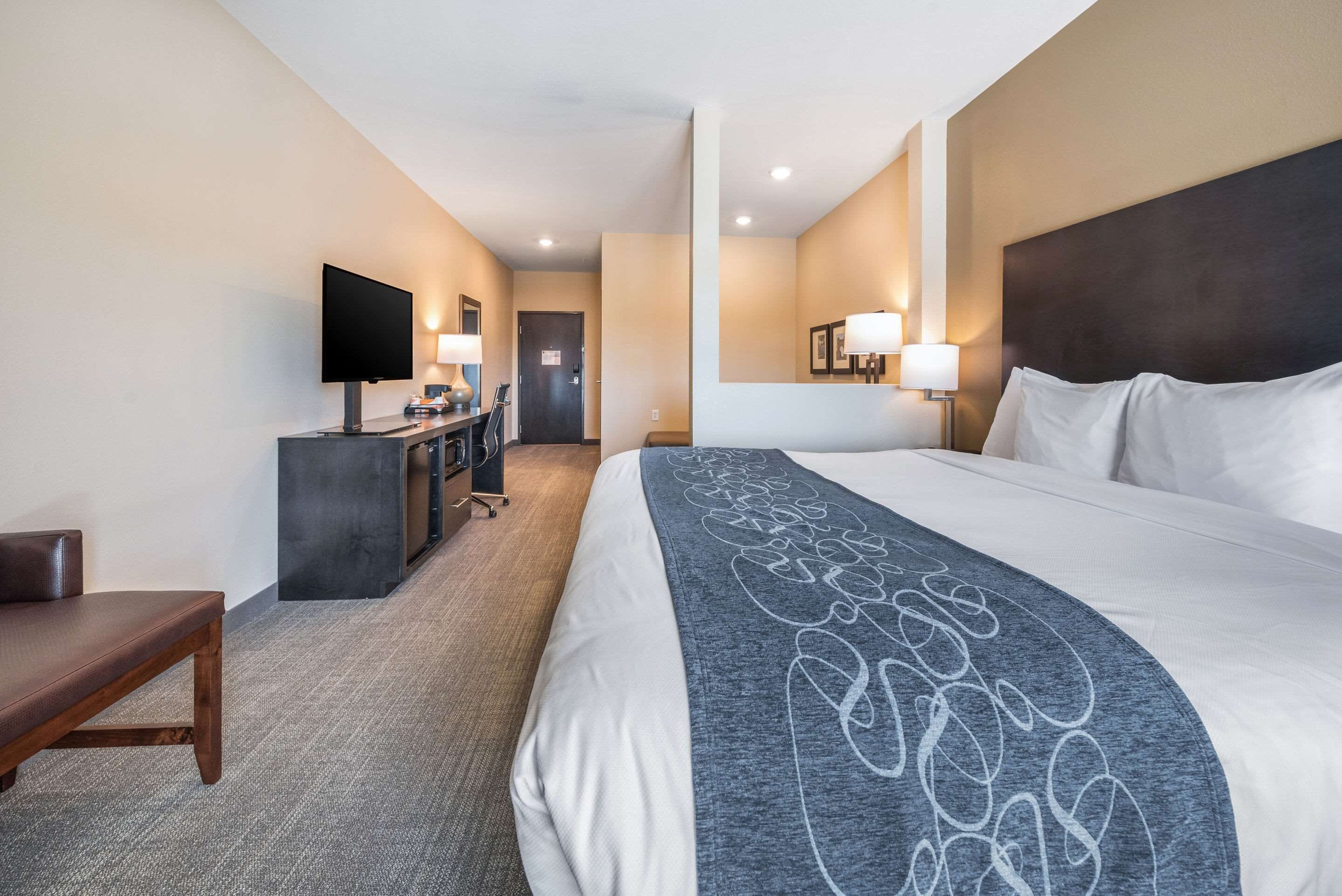 A hotel room with a large bed, a desk with a flat-screen TV, a bench, and a door leading to a hallway.