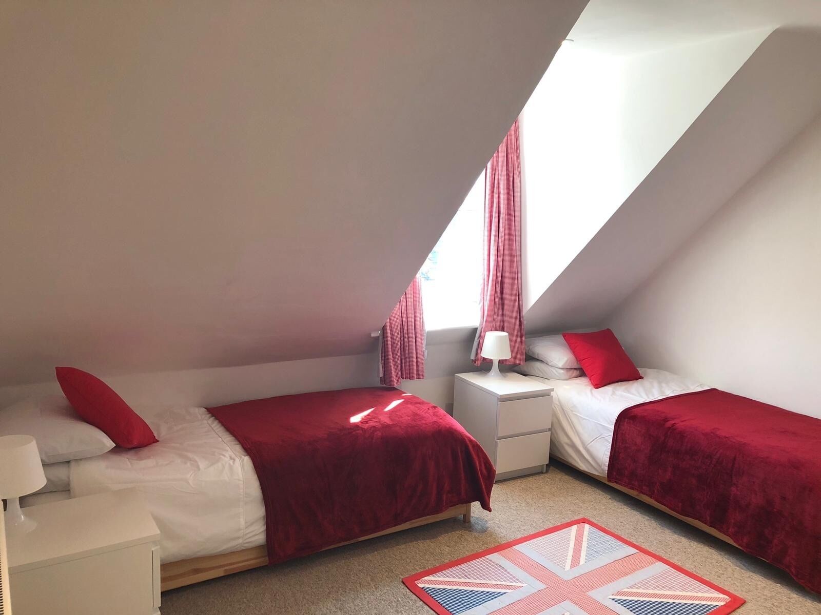 A bedroom with two beds, each with a red bedspread and pillows, a nightstand with a lamp, and a window with curtains.