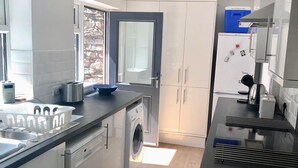 Une cuisine moderne avec des meubles blancs, un lave-linge encastré, un plan de travail noir et une fenêtre donnant sur un mur en pierre.