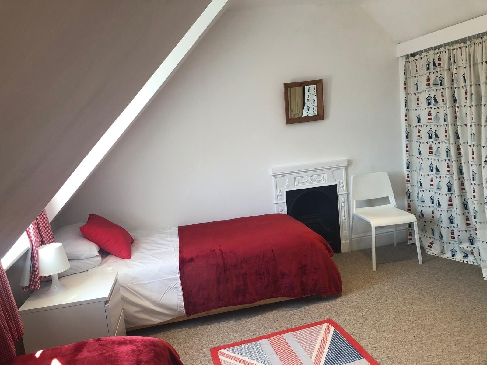 A bedroom with a sloped ceiling, a bed with red bedding, a white chair, a fireplace, and a patterned curtain.