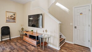 Een woonkamer met een televisie, een glazen tafel, een stoel, een kleine plant en een trap.