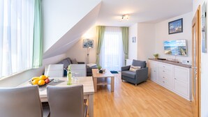A modern living room with a dining area, a sofa, a coffee table, and a kitchen area with a cabinet and a TV. - Hus Günter - Apartment Maritim (Seebad Ahlbeck)