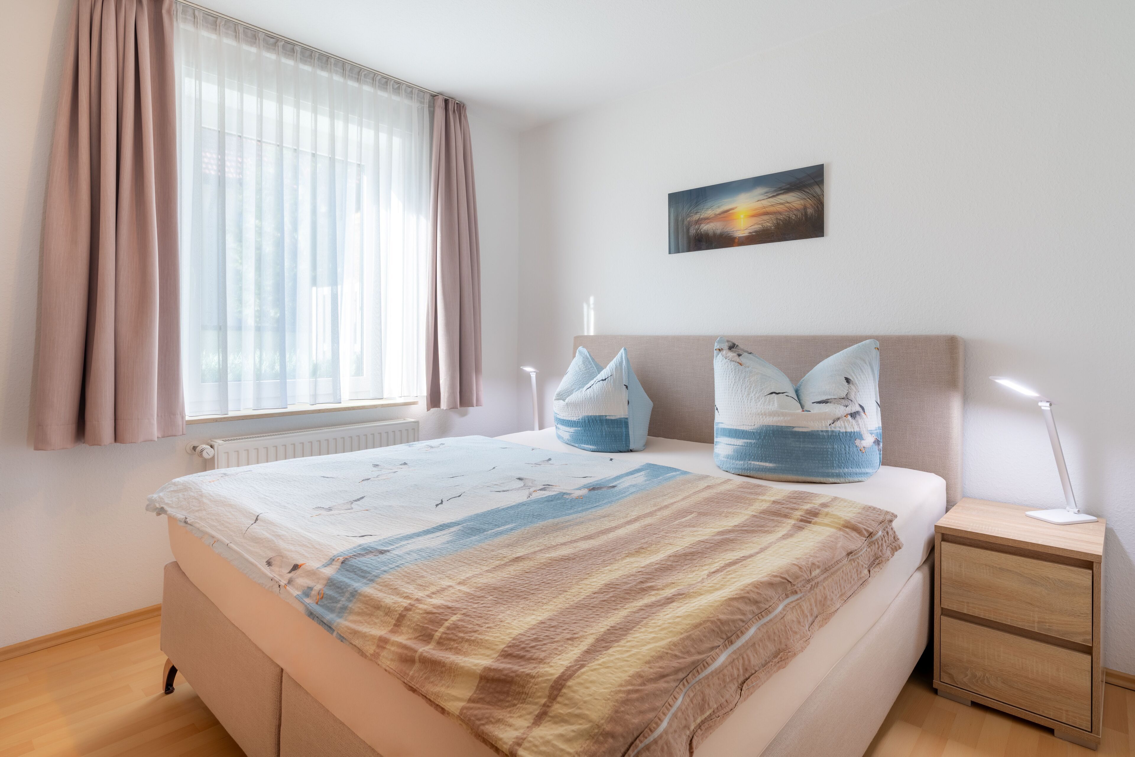 Ein Schlafzimmer mit Bett, Nachttisch, Fenster mit Vorhängen und einem Wandbild.