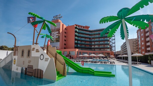 A swimming pool with a pirate-themed slide and palm trees, surrounded by a hotel building with balconies and outdoor seating. - Hotel Fenals Garden (Lloret de Mar)