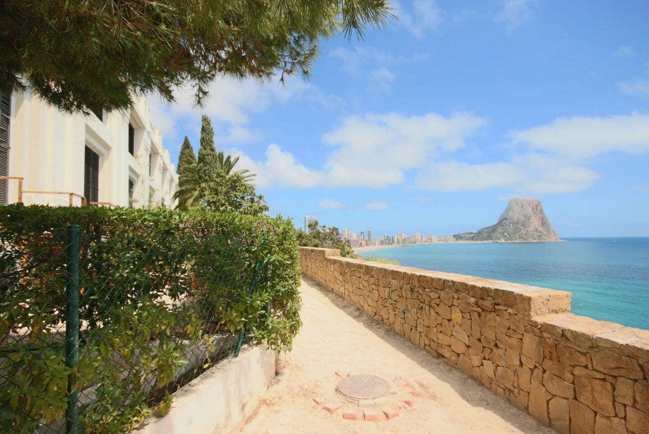 A coastal walkway with a stone wall, a clear view of the sea, and a distant mountain.