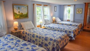 Une chambre avec trois lits, chacun recouvert d’une couvre-lit à motifs floraux bleus et blancs. Il y a deux fenêtres avec des rideaux blancs, un tableau représentant un phare sur le mur, et une petite table avec une lampe placée entre les lits.