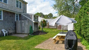 Un jardin avec une terrasse en bois, une table de pique-nique, un barbecue, un parasol et une clôture en bois.