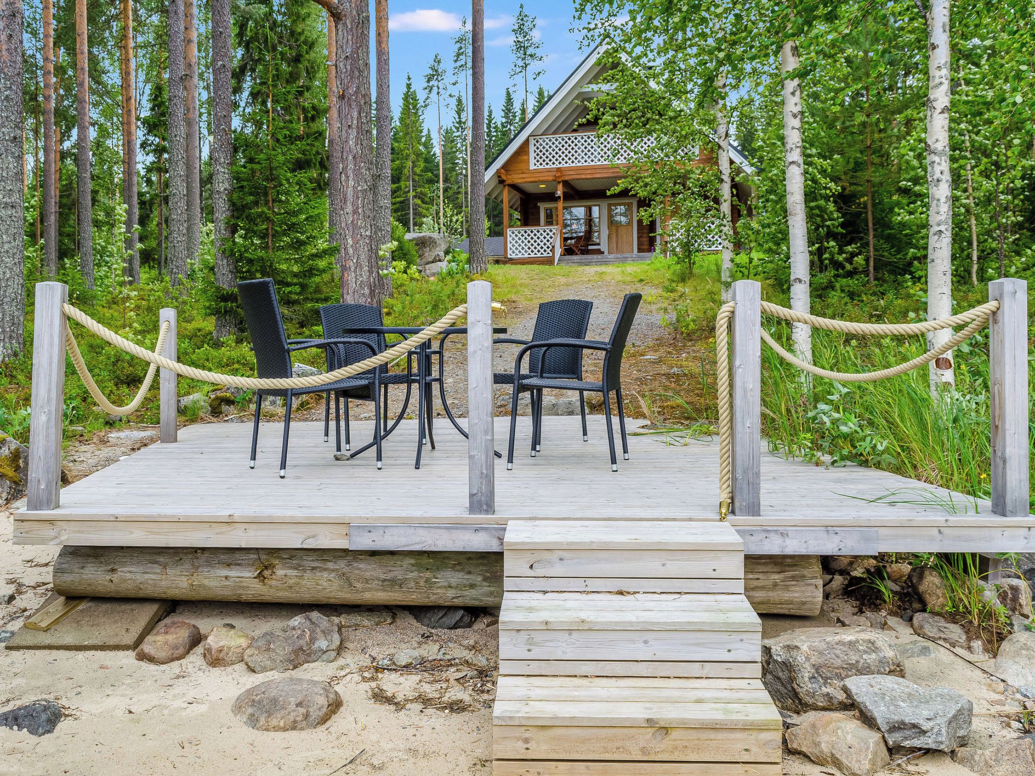 Une terrasse en bois, dotée de chaises et d’une barrière en corde, mène à un chalet dont la façade est ornée d’un treillis.