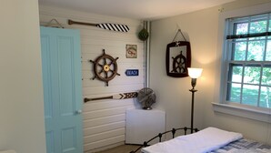 Ein Schlafzimmer mit einem Bett, einer Holztür, einem Wandventilator, einem Schiffsschwungrad und einem Fenster mit Blick auf Grünfläche.
