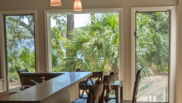 A kitchen with a central island, wooden bar stools, pendant lighting, and large windows offering views of greenery. - 3 Bdrm 2 Bath Waterfront Villa on Dataw with Dock, Bikes, Pool and Beach Pass!  (St Helena Island)