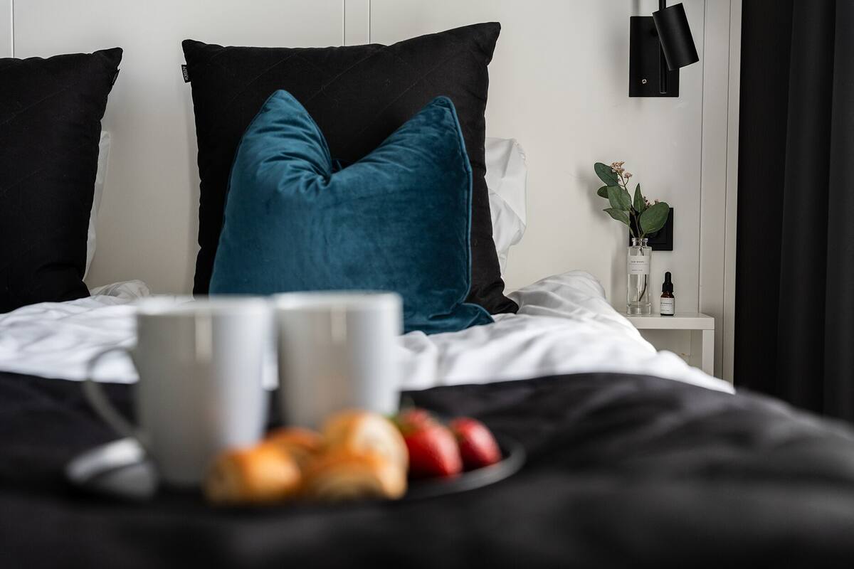 Un lit bien fait, avec deux oreillers, une table de chevet ornée d’un vase et d’une bouteille, et une assiette de fruits.