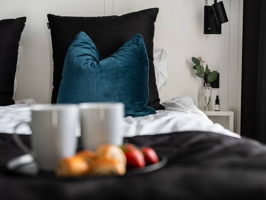 A neatly made bed with two pillows, a bedside table with a vase and a bottle, and a plate of fruit.