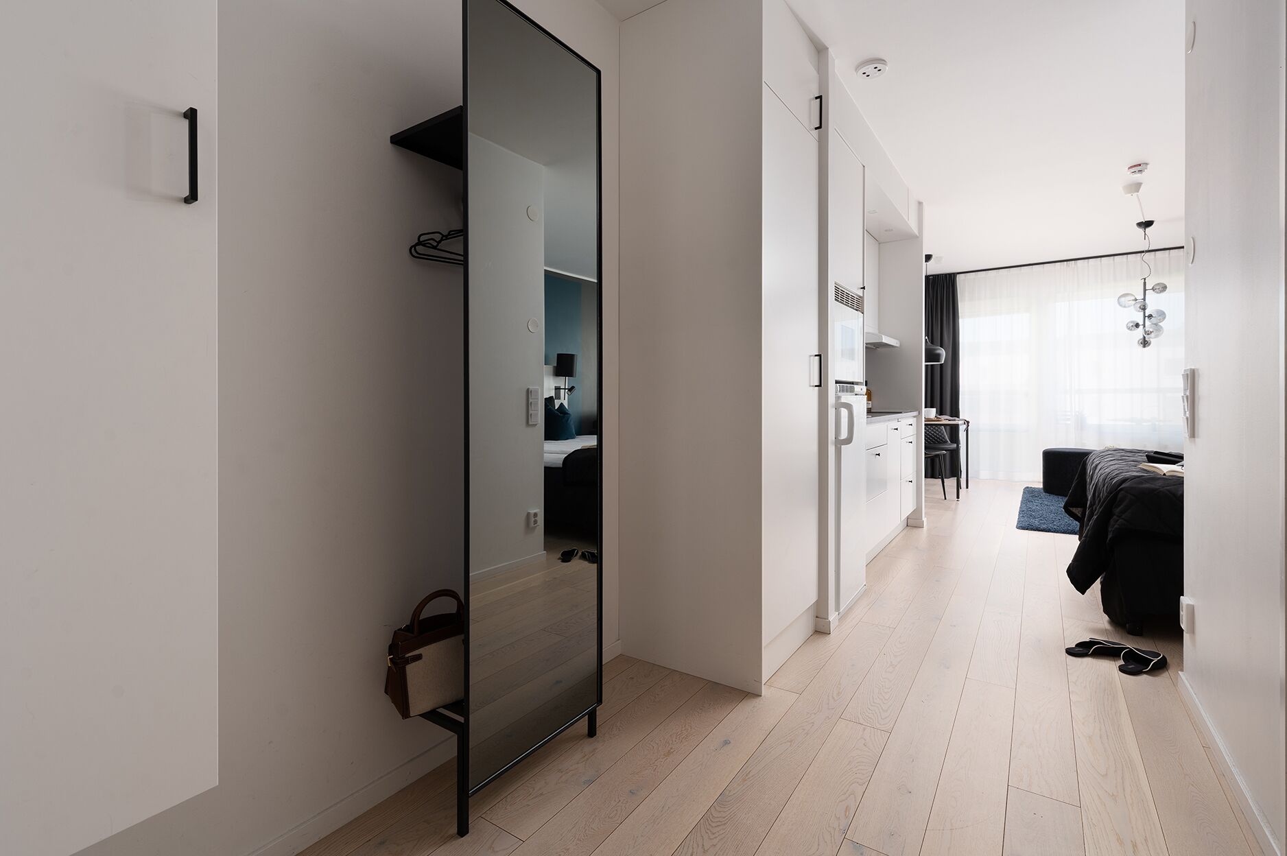 A modern hotel room with a glass partition, a kitchenette, a seating area with a sofa and a small table, and a view of the city through large windows.