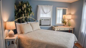 Een slaapkamer met een bed, nachtkastjes, een spiegel, een raam met gordijnen en een wandairconditioner.