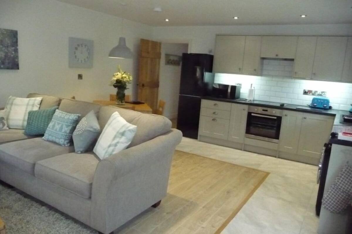 A modern living room with a beige sofa, a wooden coffee table, and a kitchen area with light-colored cabinets and appliances.