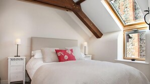 Una camera da letto con soffitto spiovente, una finestra a lucernario, un letto con biancheria da letto bianca e un cuscino rosso, due comodini con lampade e vista su un edificio in mattoni dalla finestra.