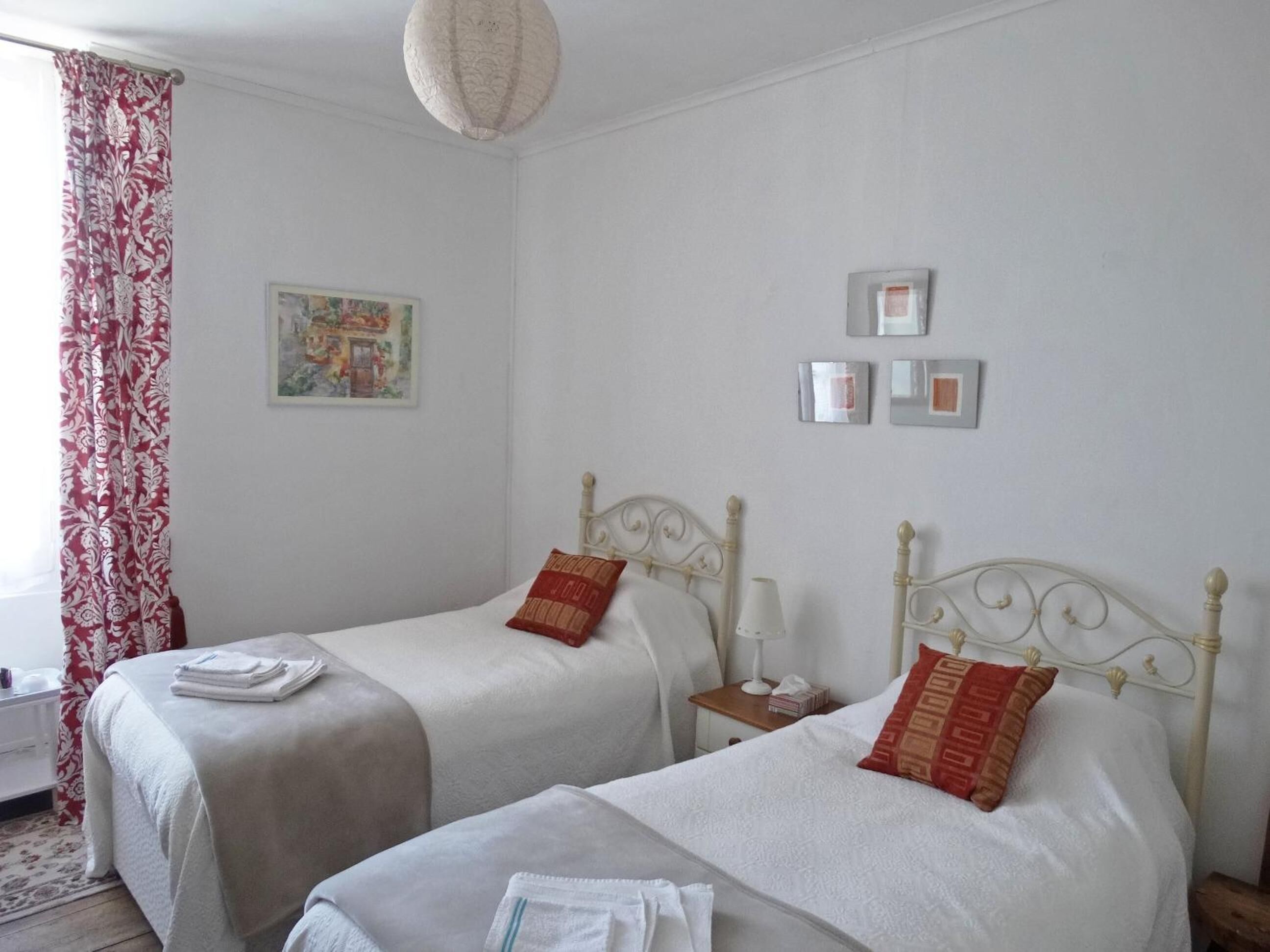 Une chambre double avec deux lits, une petite table de chevet avec une lampe, un tableau encadré au mur et une fenêtre avec des rideaux rouges.