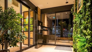 Entrada de un hotel con puerta de cristal, plantas de interior y un cartel que dice 'SuperHotel'.