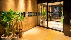 Entrada de un hotel con paneles de madera, un letrero que dice SUPERHOTEL y plantas en macetas.