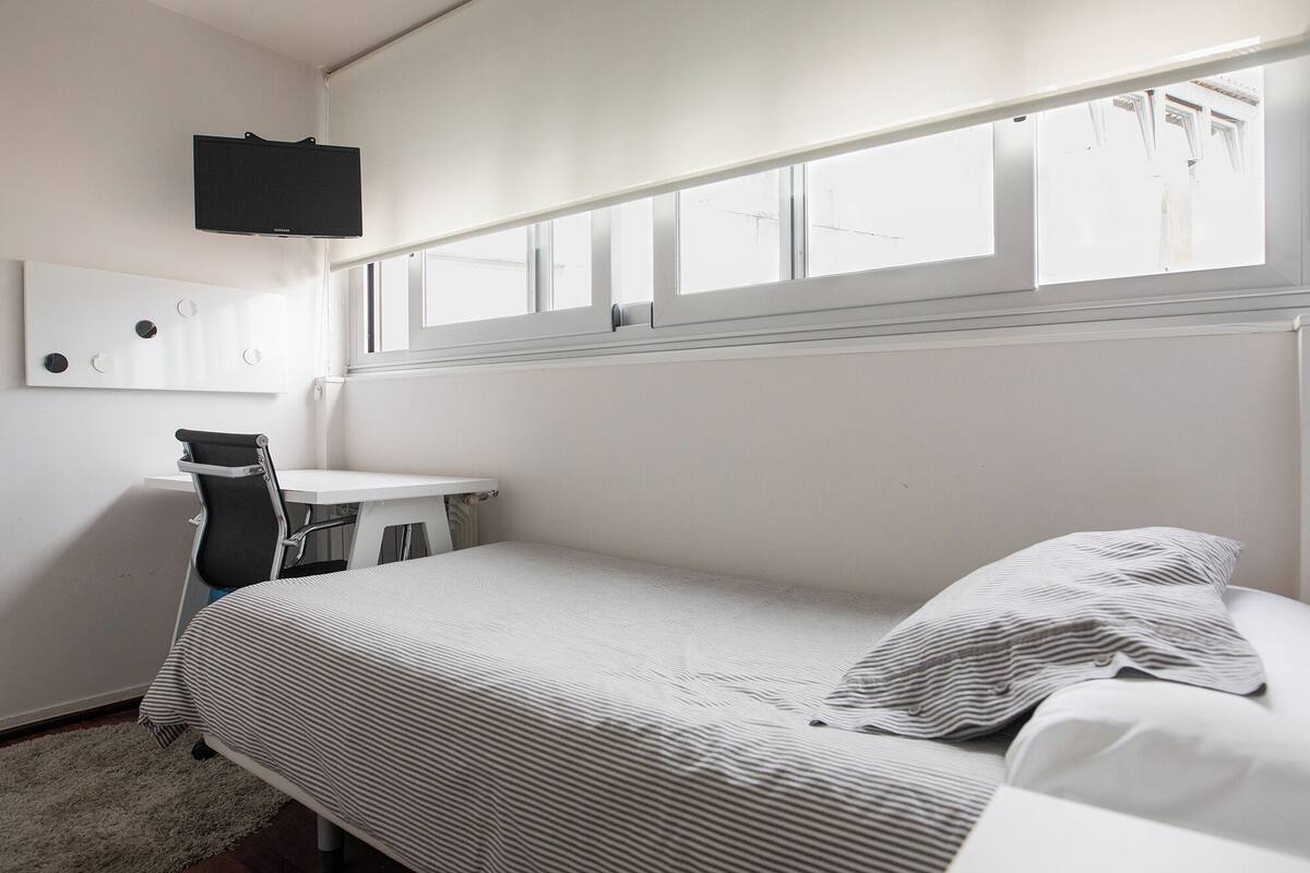 Een kamer met een enkel bed, een raam, een bureau en een stoel.