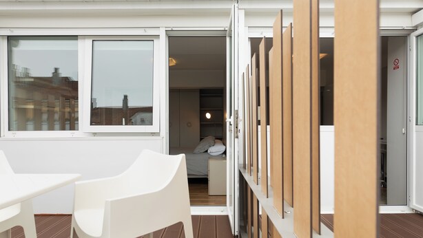 Een moderne kamer met een bed, een klein tafeltje en een stoel. Er is een raam met uitzicht op gebouwen.