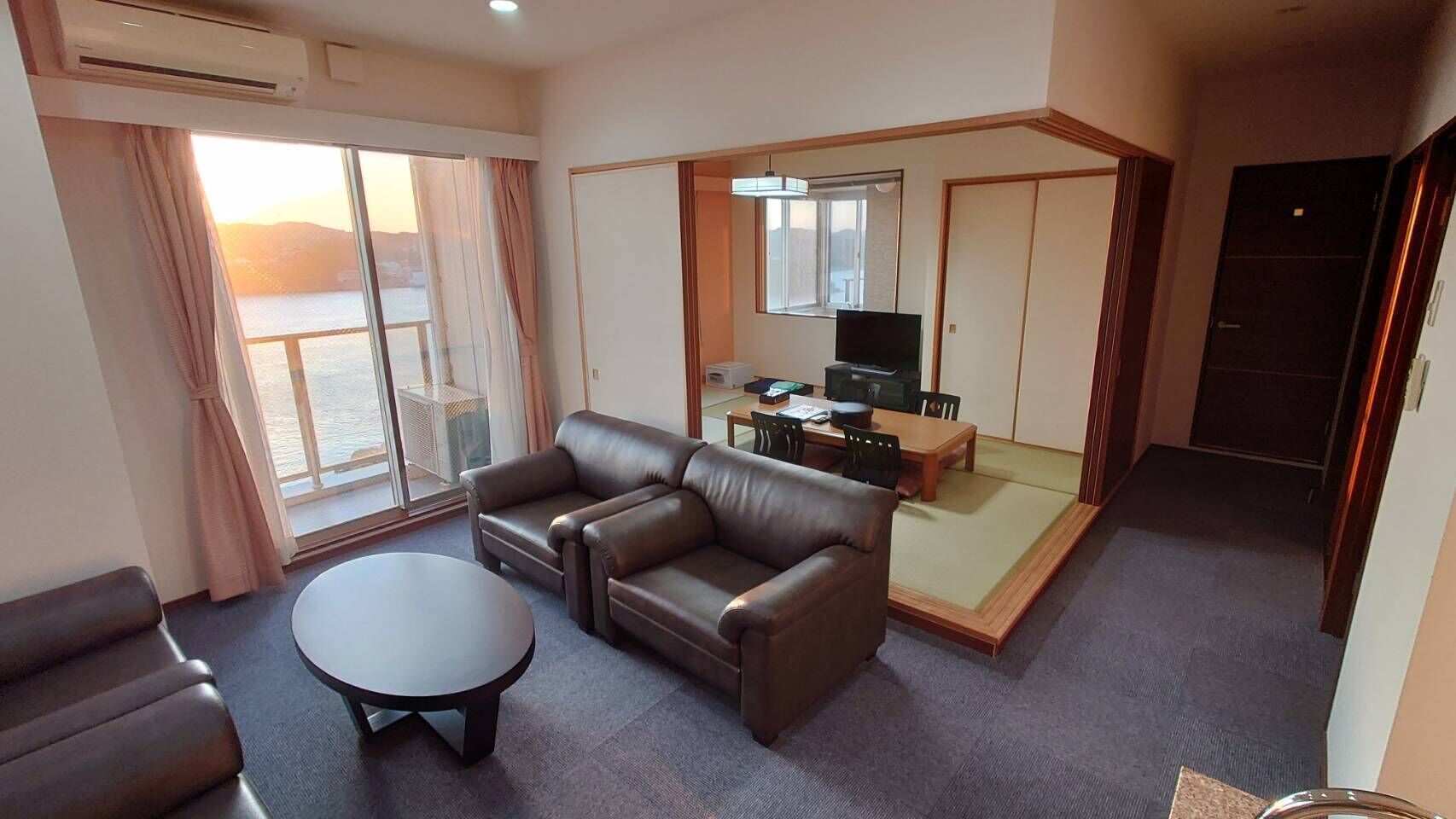 A hotel room with a sofa, a small table, a TV, and a sliding door leading to a balcony with a view.