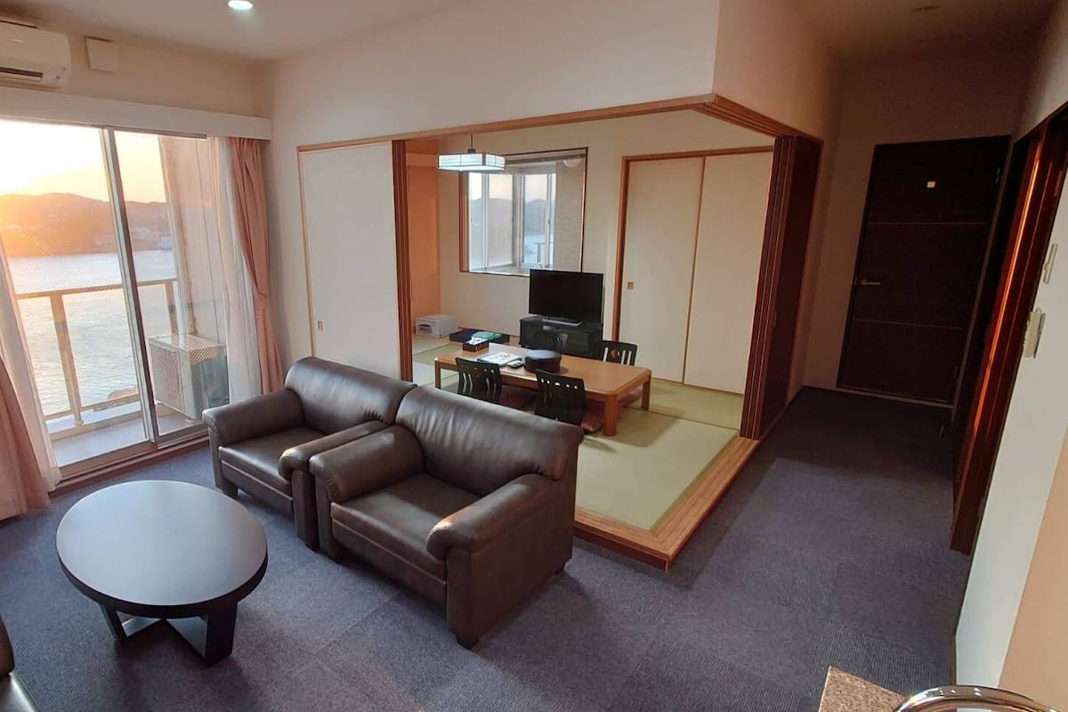 Habitación de hotel con sofá, una mesita, televisor y puerta corredera que da a un balcón con vistas.