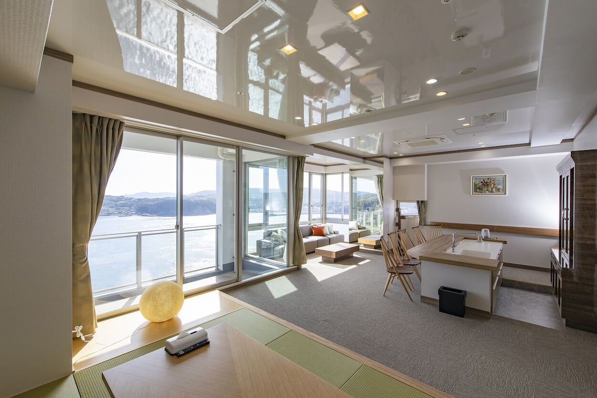 Una habitación de hotel moderna con un gran ventanal corredero a un balcón, una zona de estar con sofá y mesa de centro, una pequeña cocina y vistas al mar.